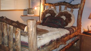 A rustic bed with a wooden frame and a fur throw, a television on a shelf, and a winter-themed mural on the wall.