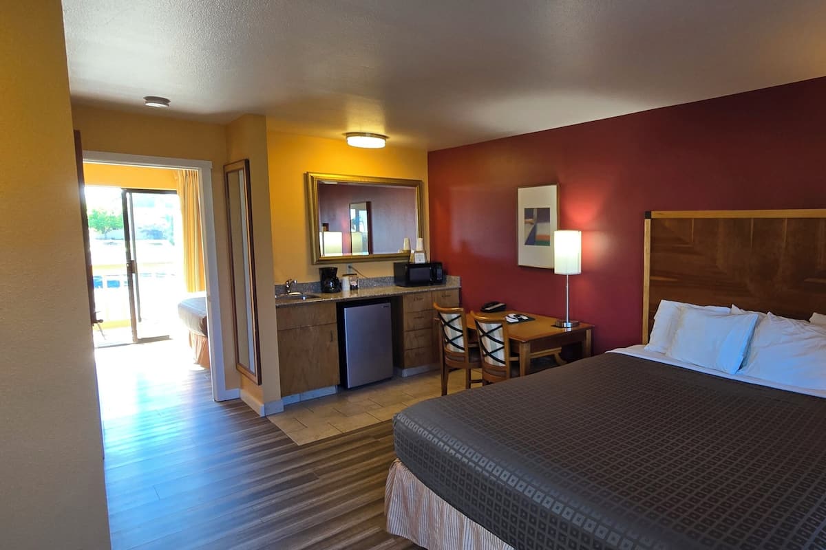Quarto de hotel com uma cama, uma secretária com cadeira, uma cozinha compacta e uma janela grande.