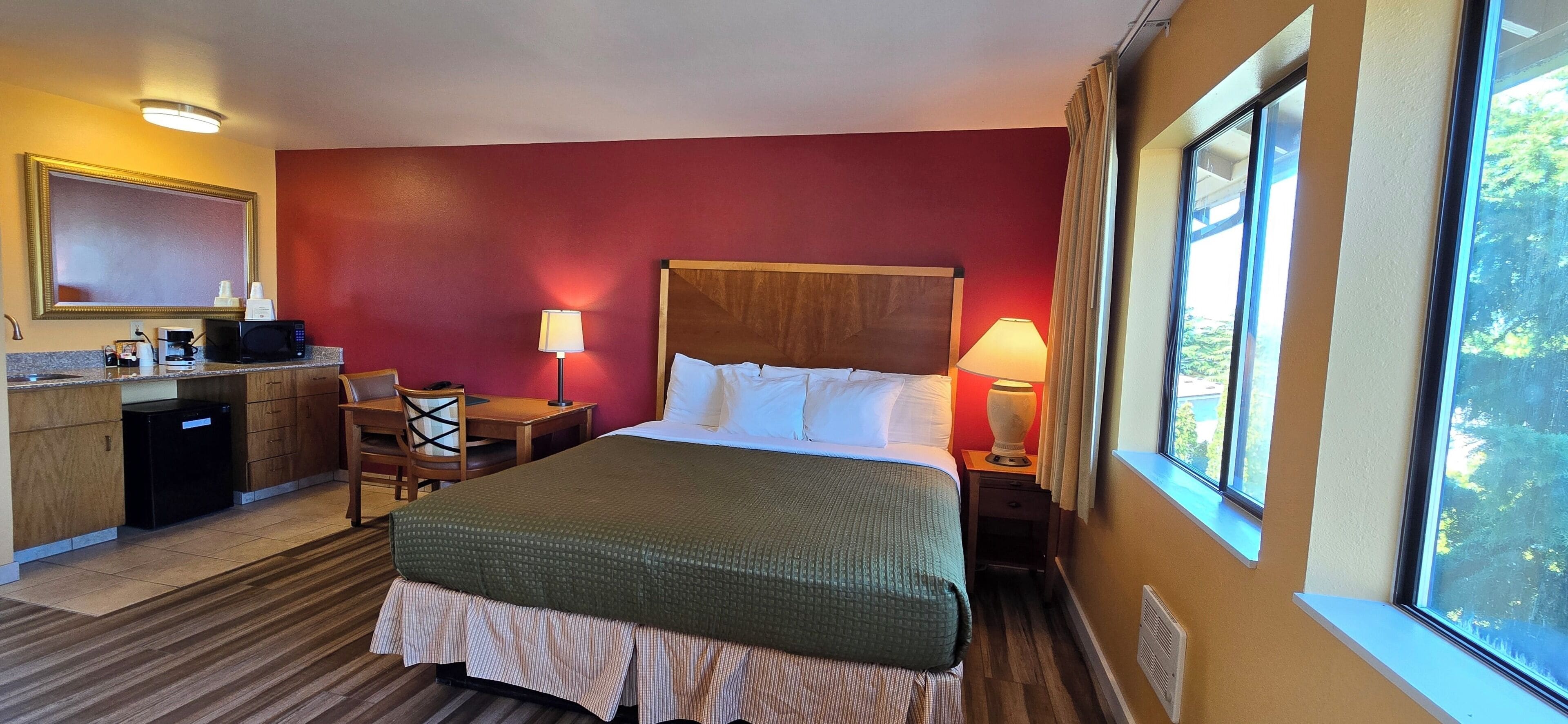 A hotel room with a bed, bedside table, lamp, desk with a chair, and a small kitchenette.