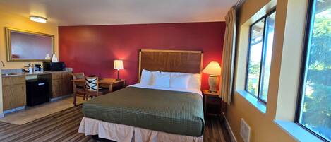 A hotel room with a bed, bedside table, lamp, desk with a chair, and a small kitchenette.