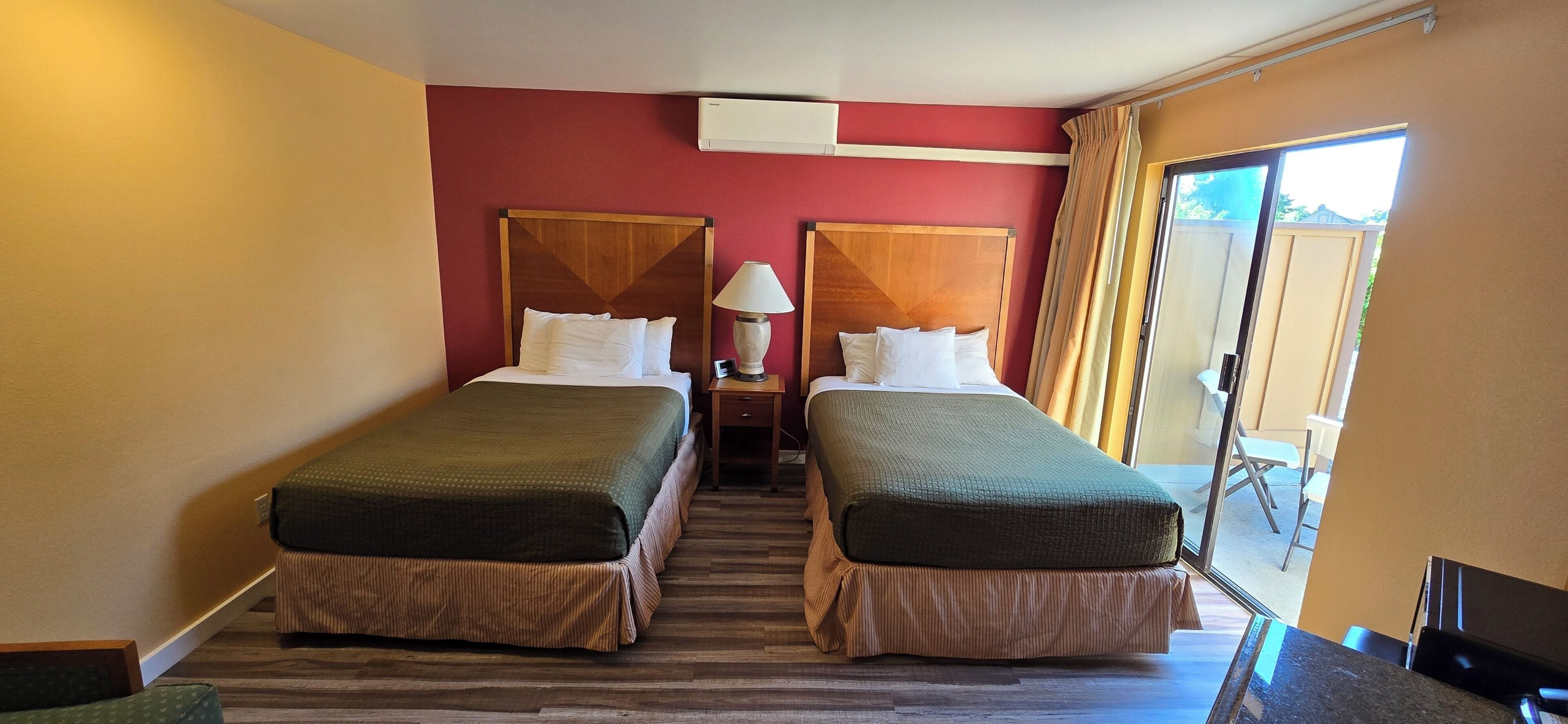 A hotel room with two beds, a nightstand, a lamp, and a sliding door to a balcony.