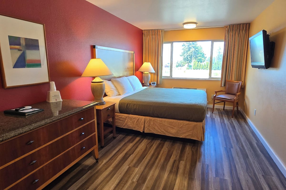 Quarto de hotel com cama, luminárias laterais, escrivaninha com telefone, cadeira e televisão fixada na parede.