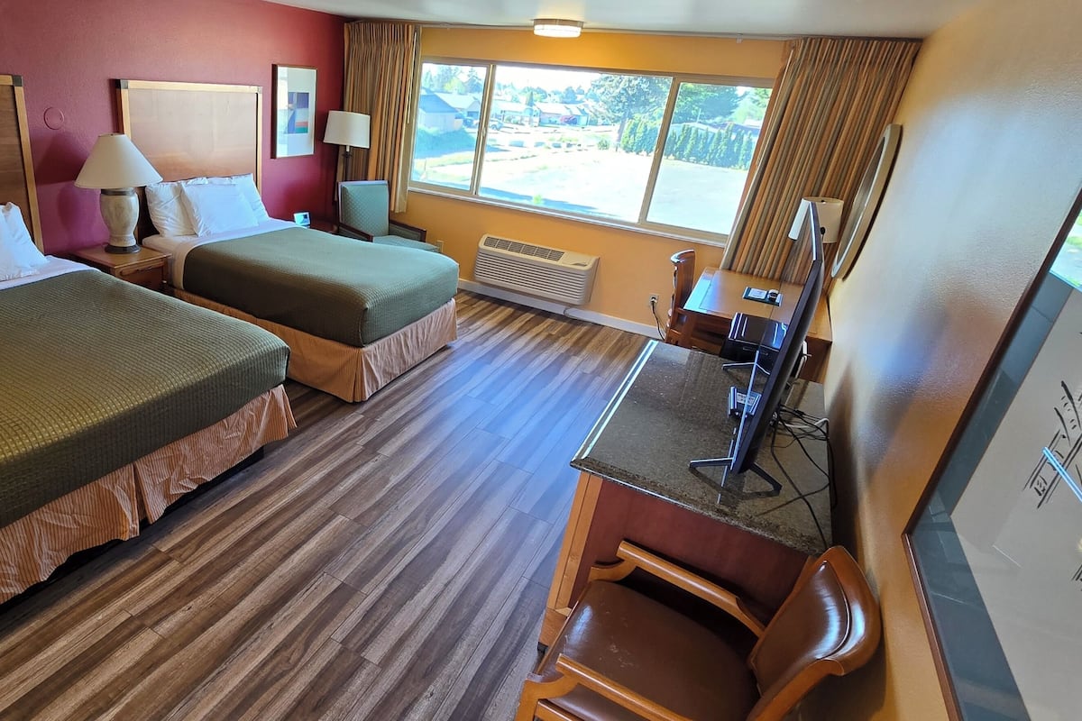 Quarto de hotel com duas camas, uma escrivaninha, uma cadeira, uma janela com vista e um quadro na parede.