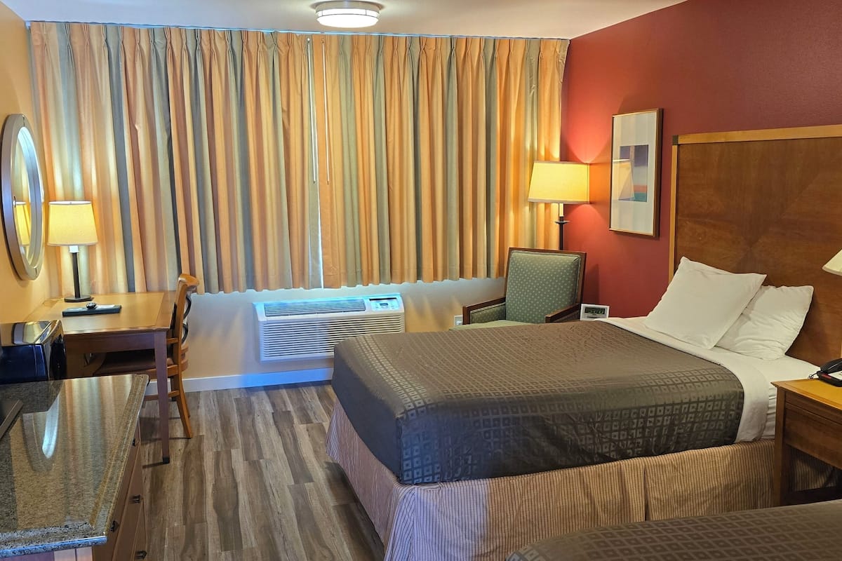 Quarto de hotel com cama, mesa de cabeceira, luminária, televisão e cortinas.