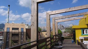 Een dakterras met houten reling, een geel gebouw en uitzicht op een stadsgezicht inclusief een hoge schoorsteen.