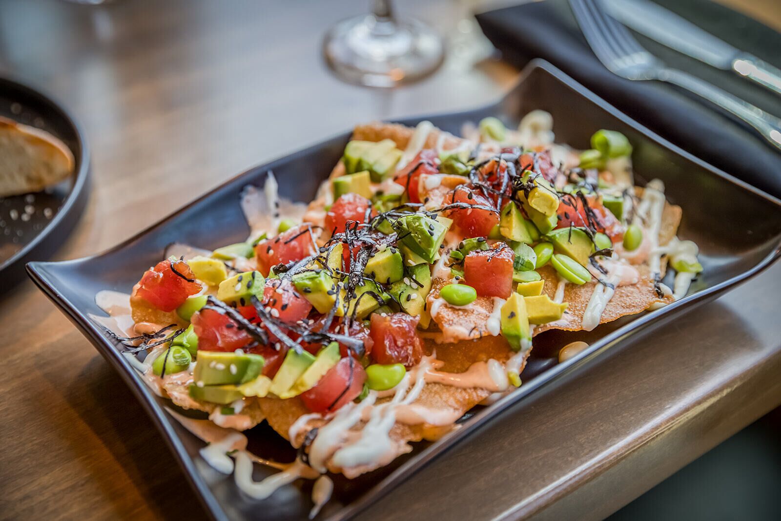 Un plato rectangular con nachos cubiertos de aguacate, tomates, guisantes verdes y una salsa cremosa.
