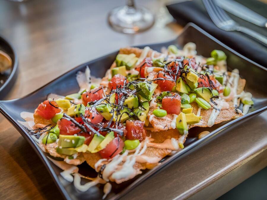 Un plato rectangular con nachos cubiertos de aguacate, tomates, guisantes verdes y una salsa cremosa.