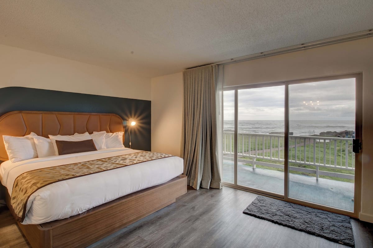 A hotel room with a large bed, a view of the ocean through a sliding glass door, and a balcony with a railing.