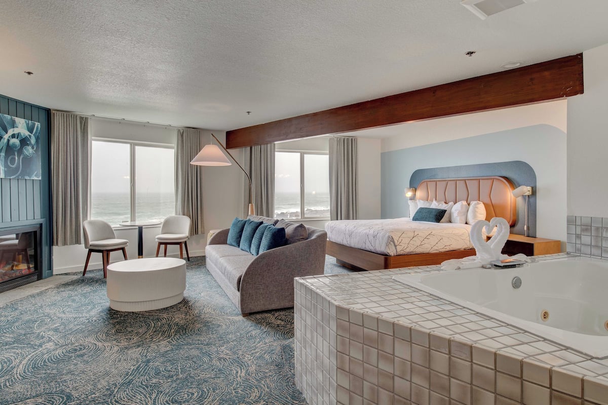 A hotel room with a large bed, a sitting area with a sofa and chairs, a small round table, a bathtub, and a view of the sea.