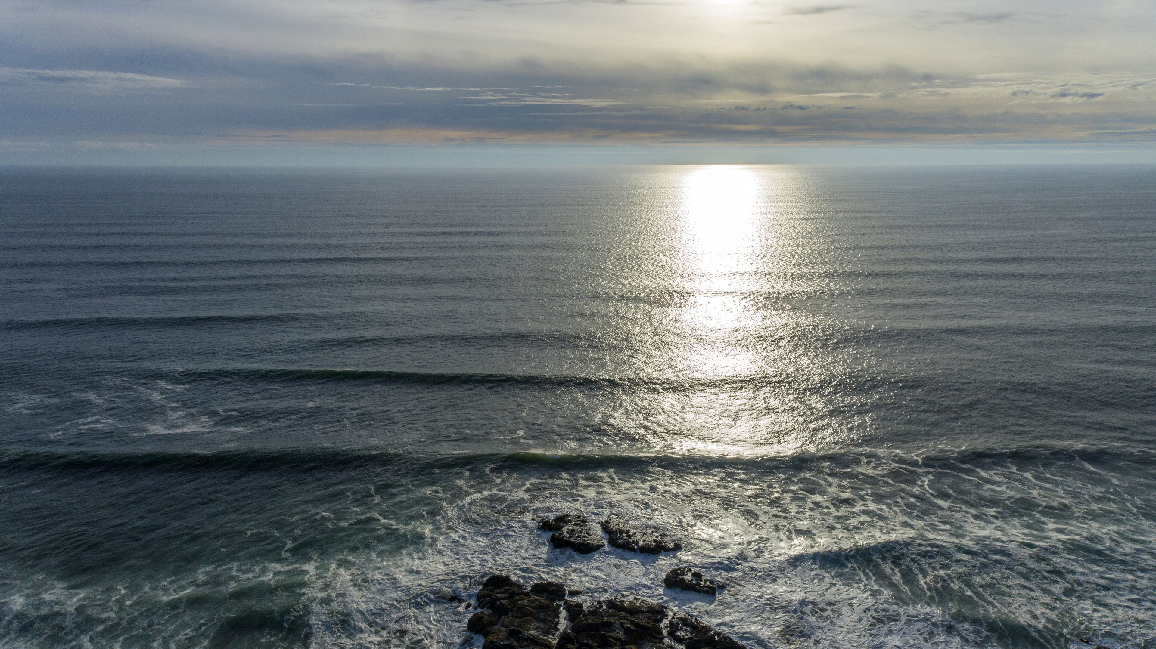 A coastal view with waves and rocks in the foreground, and the sun reflecting off the water.