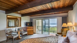 A hotel room with a large bed, a TV, a desk with a chair, a dresser, and a balcony with a view of the ocean. - Adobe Resort (Yachats)