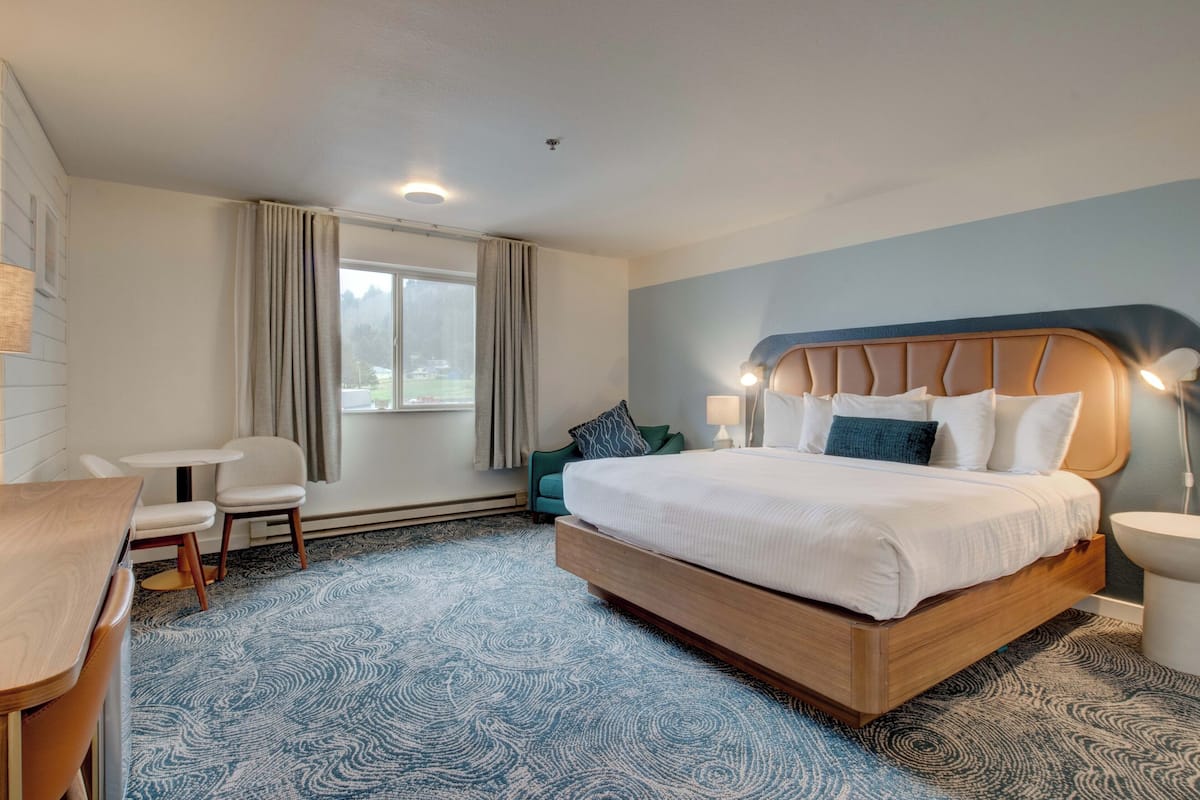 A hotel room with a large bed, a desk, and a chair. There is a window with curtains, a lamp, and a view of the outdoors.
