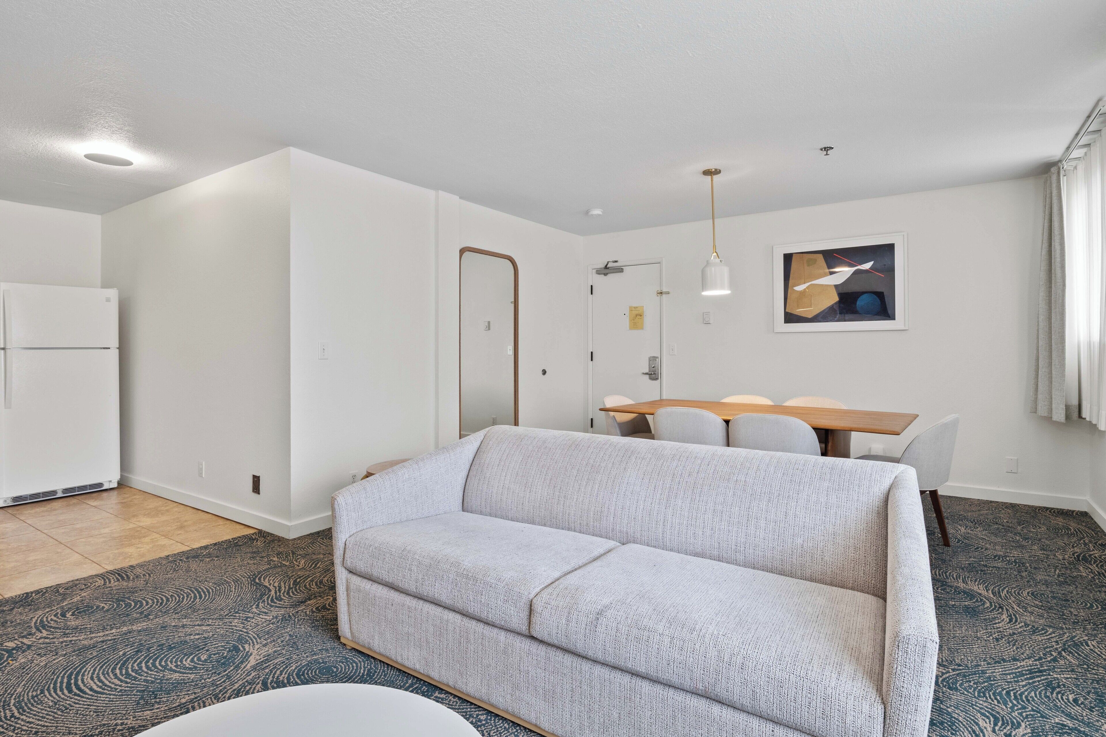 A modern living room with a sofa, a dining table with chairs, a kitchen area, and a ceiling light fixture.