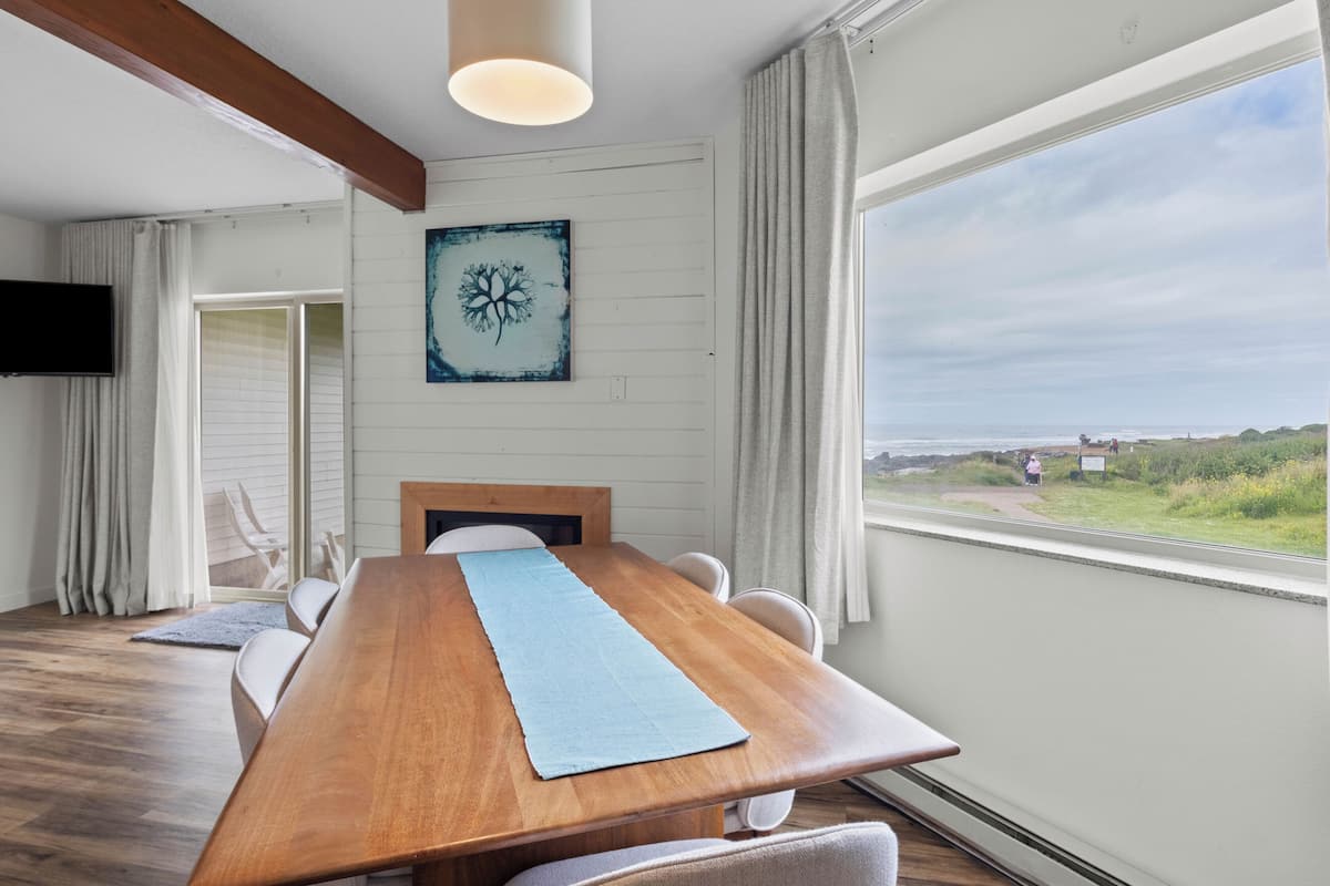 A dining area with a wooden table, chairs, a blue placemat, a mounted artwork, and a view of the beach.