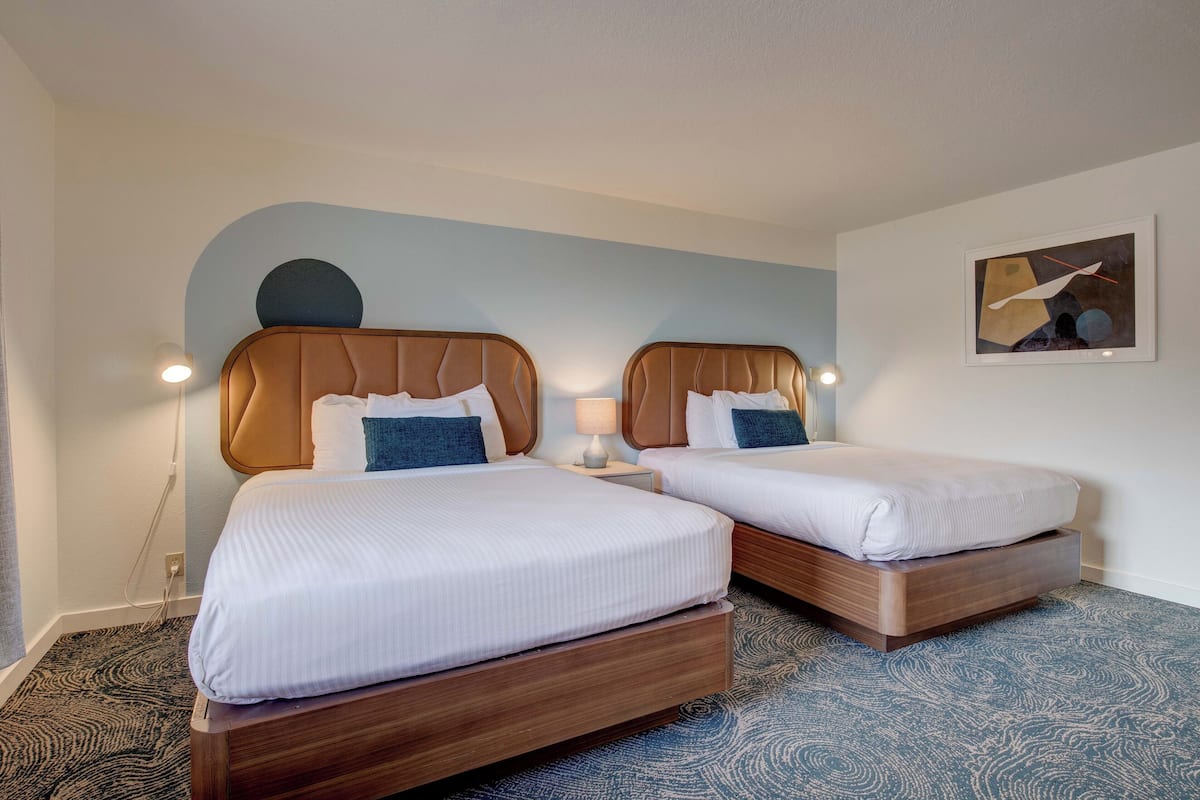 A hotel room with two beds, each with a wooden headboard and white bedding. There is a lamp on the nightstand beside each bed, and a painting on the wall.
