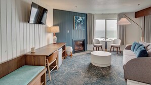 A modern hotel room with a wooden desk, a sofa, a round ottoman, a fireplace, a TV mounted on the wall, and a view of the sea through large windows. - Adobe Resort (Yachats)