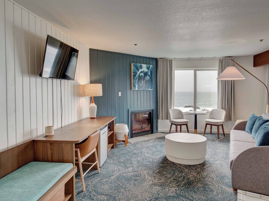 Una habitación de hotel moderna con un escritorio de madera, un sofá, un reposapiés redondo, una chimenea, un televisor montado en la pared y vistas al mar a través de amplias ventanas.