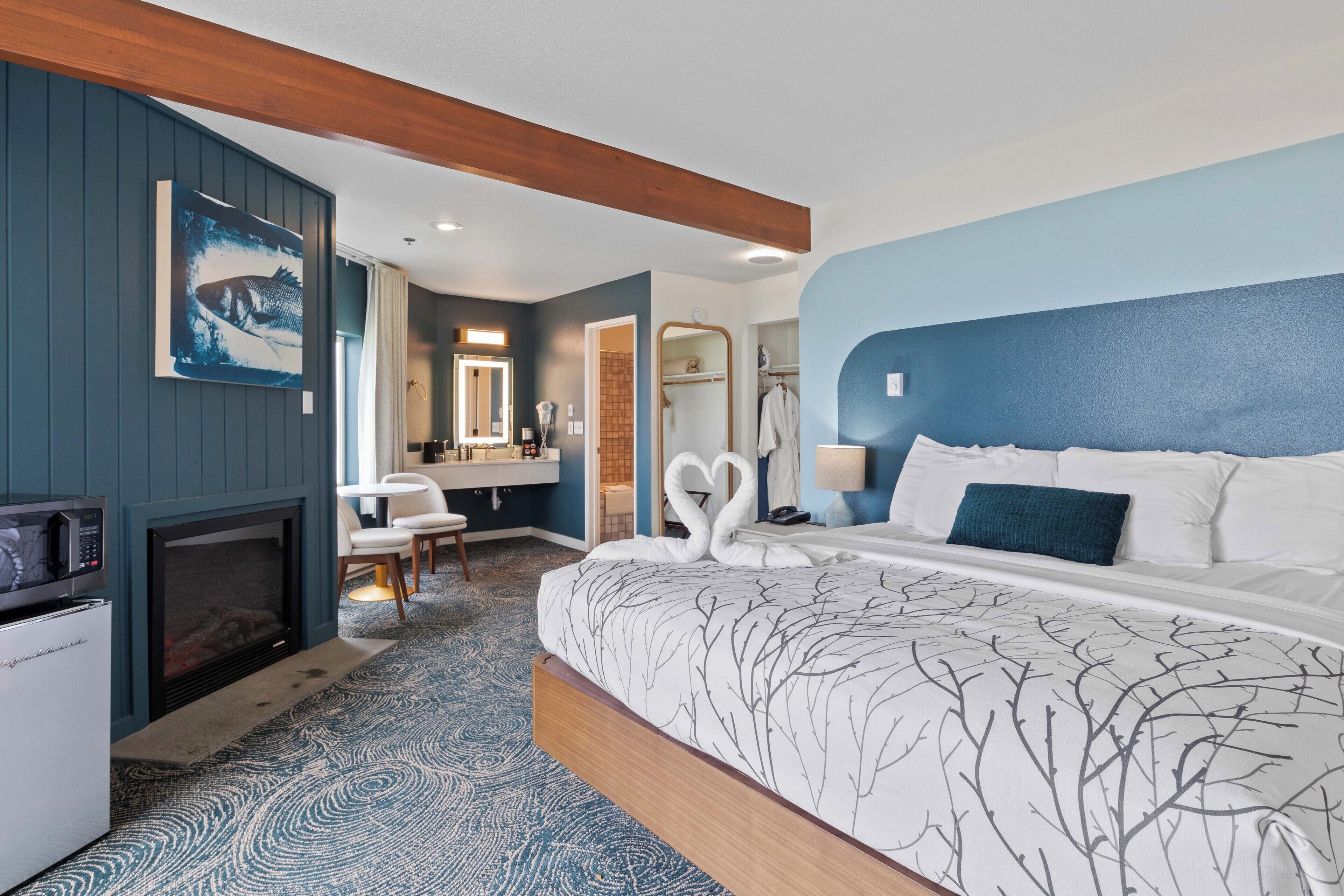 A hotel room with a large bed, a fireplace, a dining area, and a bathroom with a mirror and towel rack.
