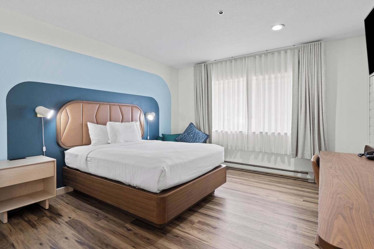 A modern hotel room with a large bed, a bedside table, a desk, and a large window with curtains.