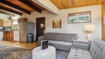 A modern living room with a grey sectional sofa, a white coffee table, and a patterned rug. There is a kitchen area with wooden cabinets and a stainless steel refrigerator.