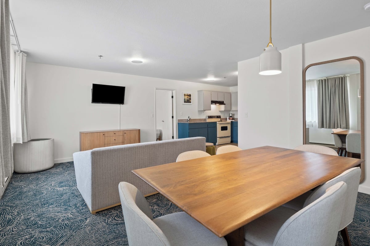 A modern hotel room with a wooden dining table, grey upholstered sofa, and a kitchen area in the background.