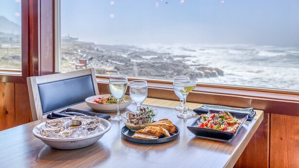 A table set with a variety of dishes and drinks, overlooking a coastal view. - Adobe Resort (Yachats)