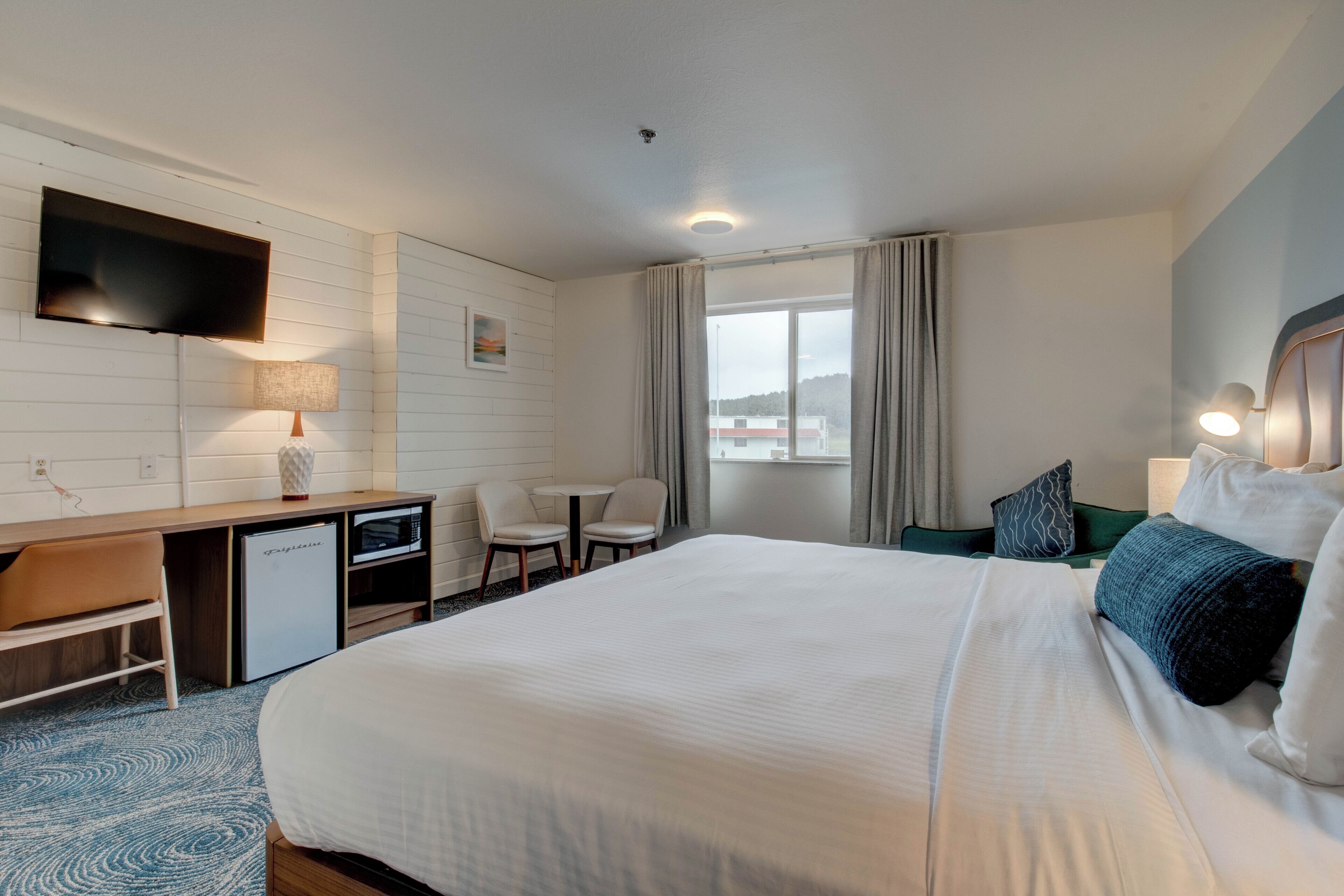 Une chambre d’hôtel avec un grand lit, un bureau avec une télévision, un petit réfrigérateur, deux chaises, une fenêtre avec des rideaux et une table de chevet avec une lampe.