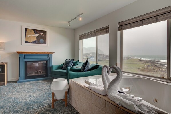 A modern bathroom with a bathtub, a large window with a view of the coast, a fireplace, and a green sofa. - Adobe Resort (Yachats)