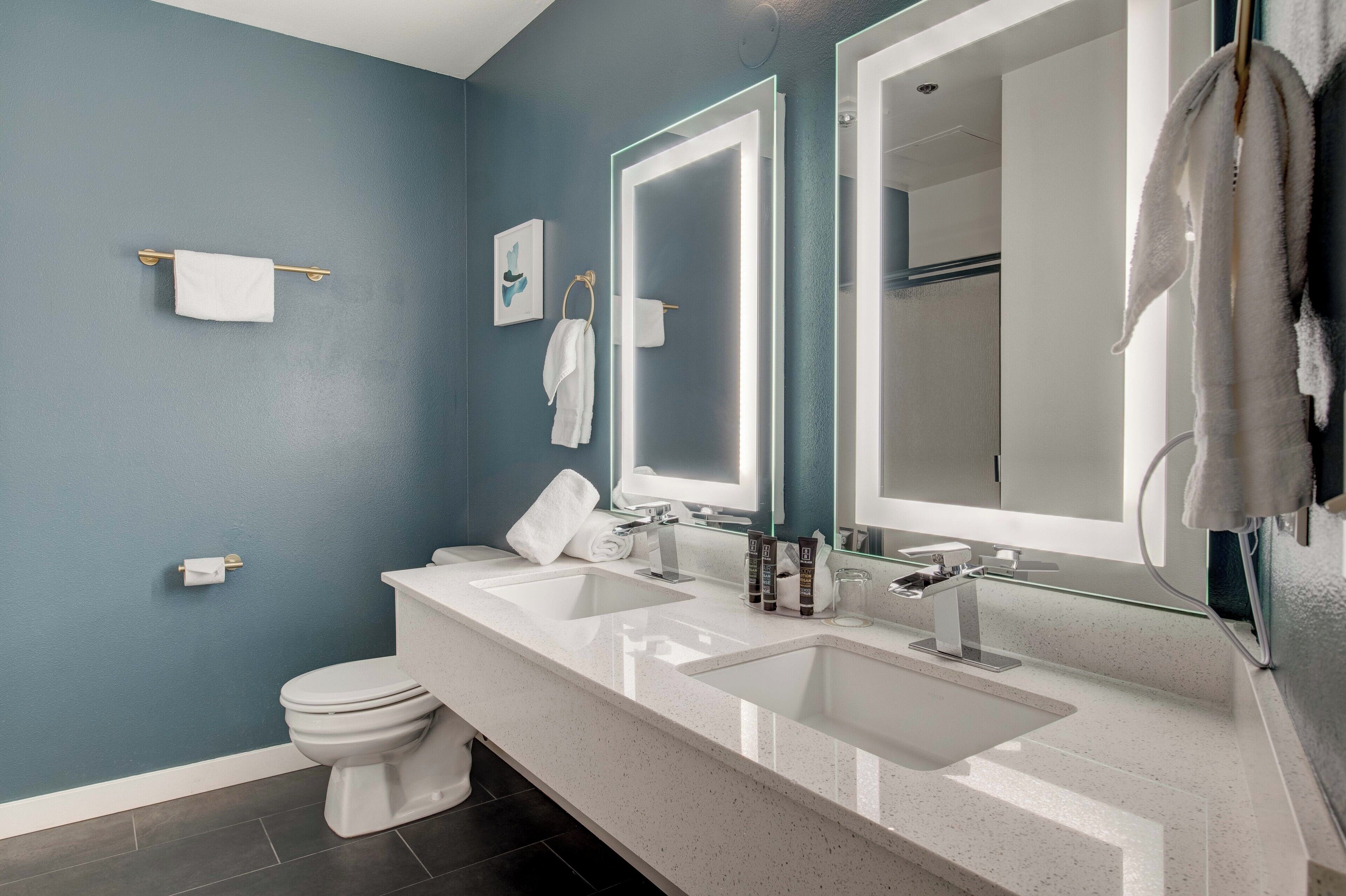 Un baño moderno con pared azul, dos lavabos, un inodoro y un espejo grande.