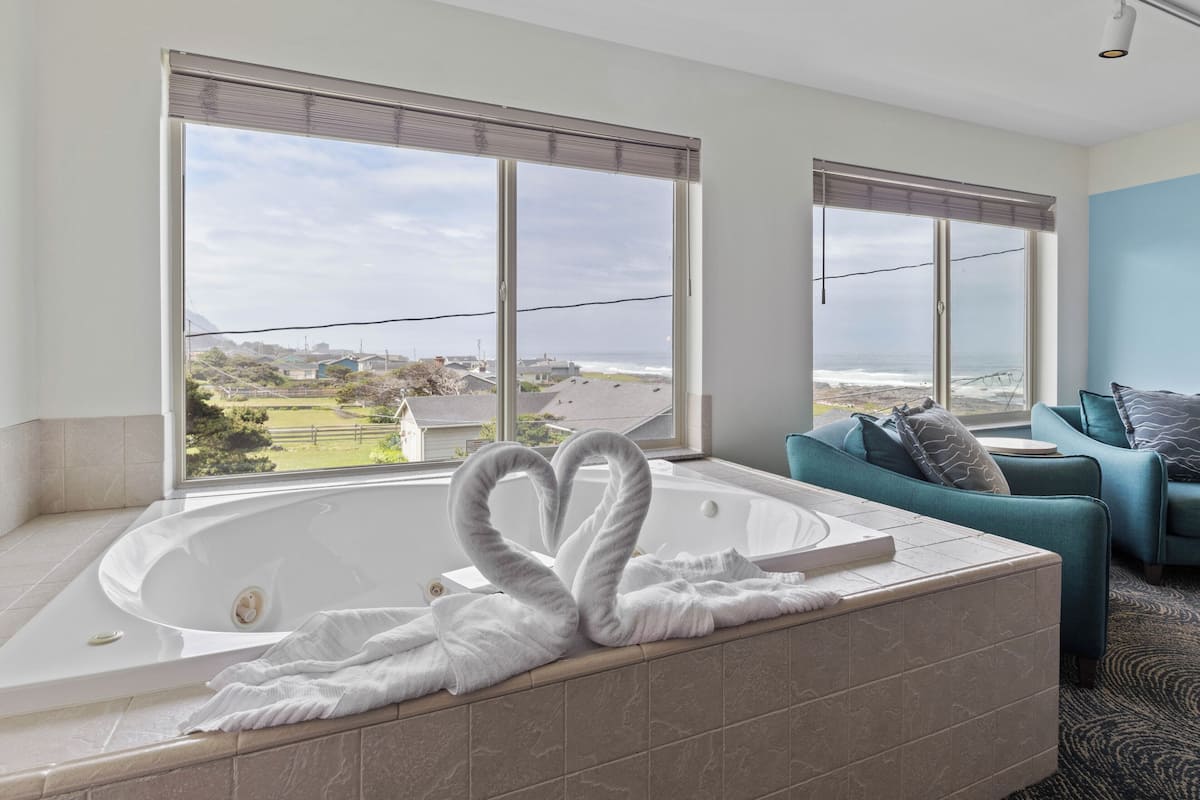 A modern bathroom with a large bathtub, a heart-shaped towel arrangement, and a view of the ocean and houses through large windows.