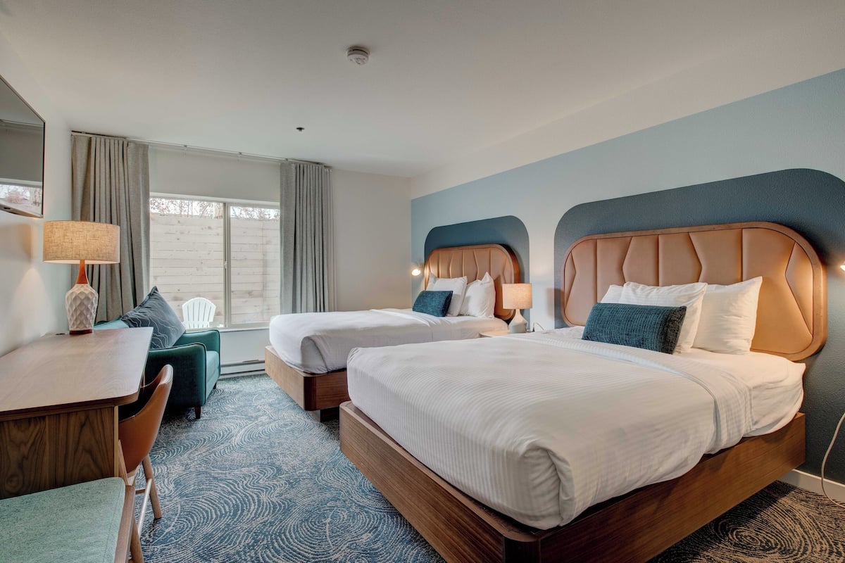 A hotel room with two beds, a desk, a chair, and a view of the outdoors through a large window.