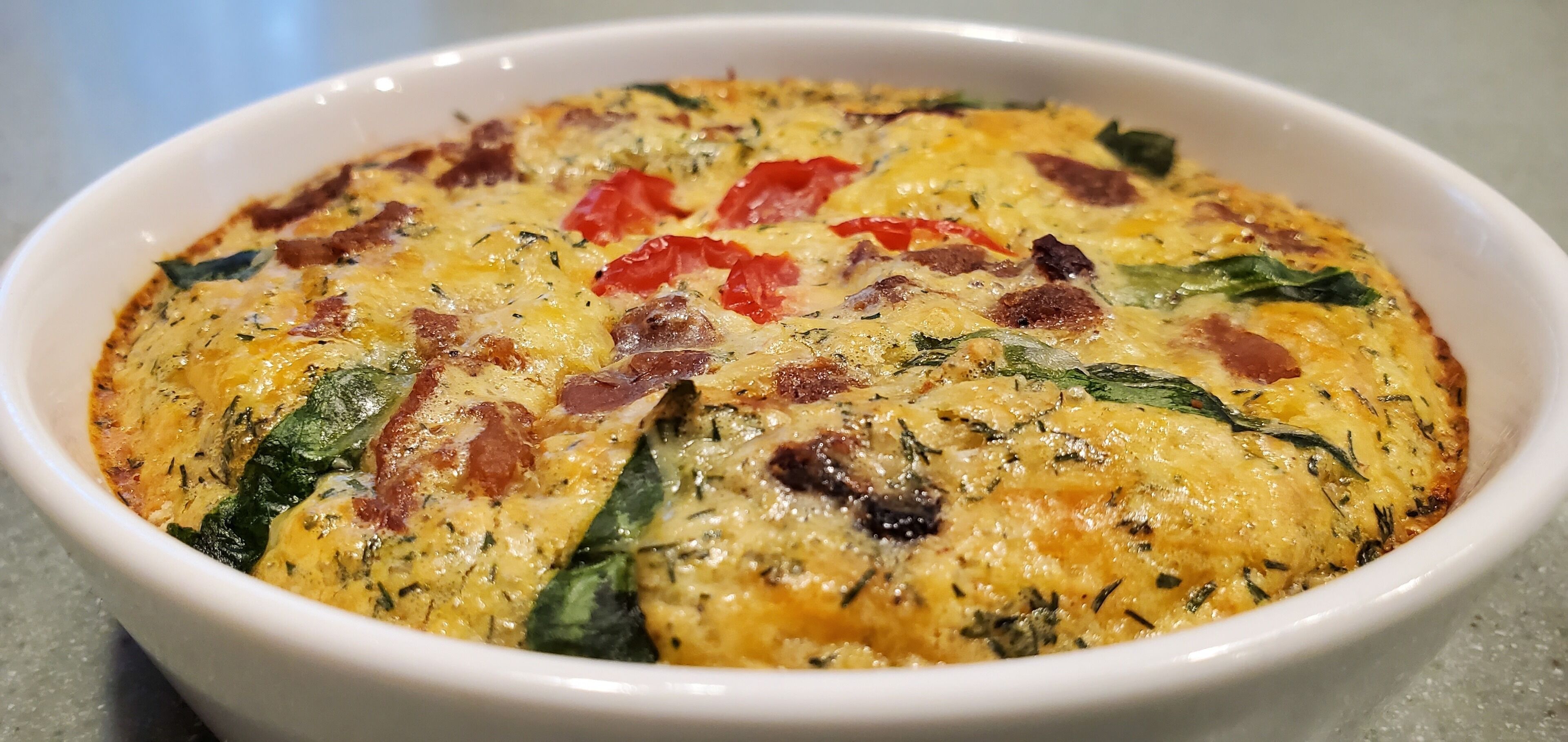 Eine weiße Schüssel mit einer gebackenen Speise, darunter Spinat, rote Paprika und Speck.