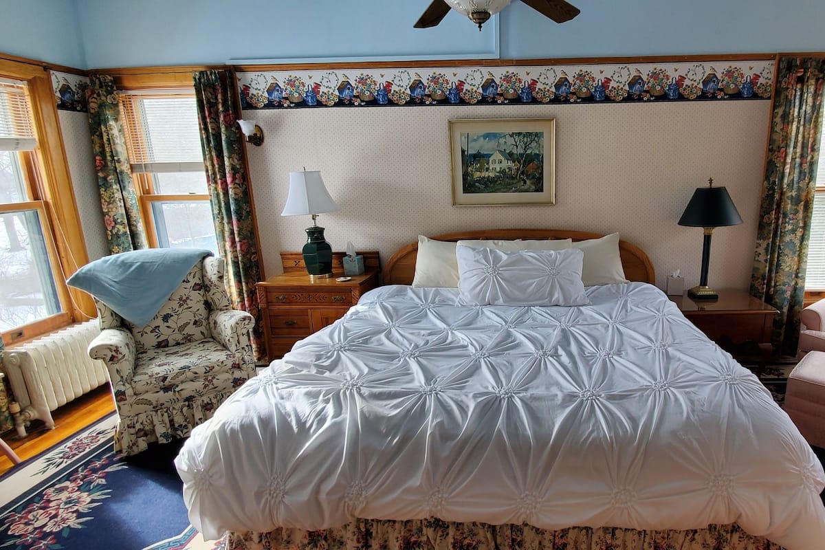 A bedroom with a large bed, two nightstands, a chair, a radiator, and a ceiling fan.