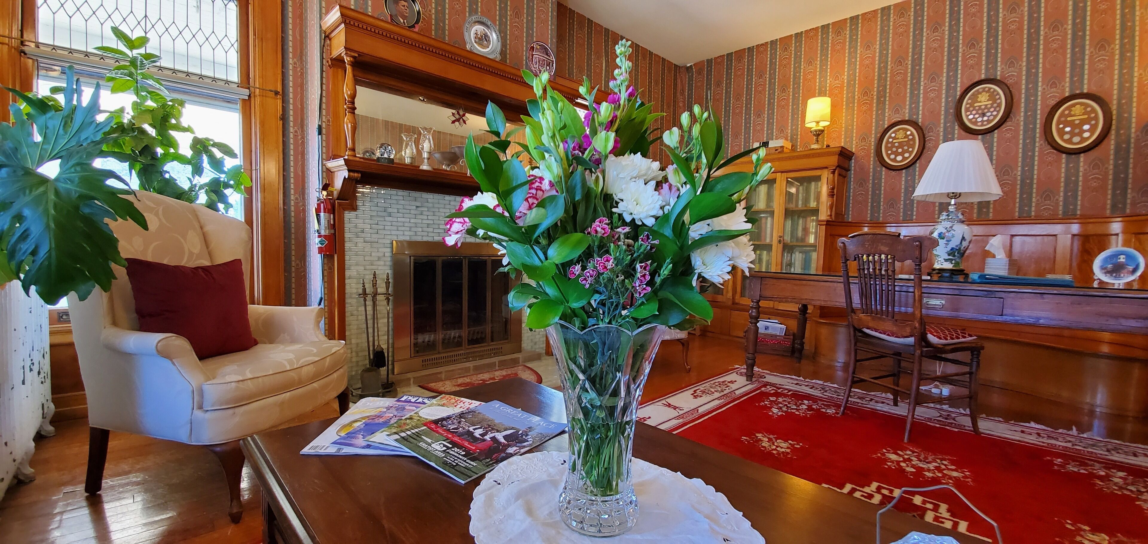 Un salón bien amoblado con chimenea, una mesa de centro con revistas, un jarrón con flores y una alfombra roja.