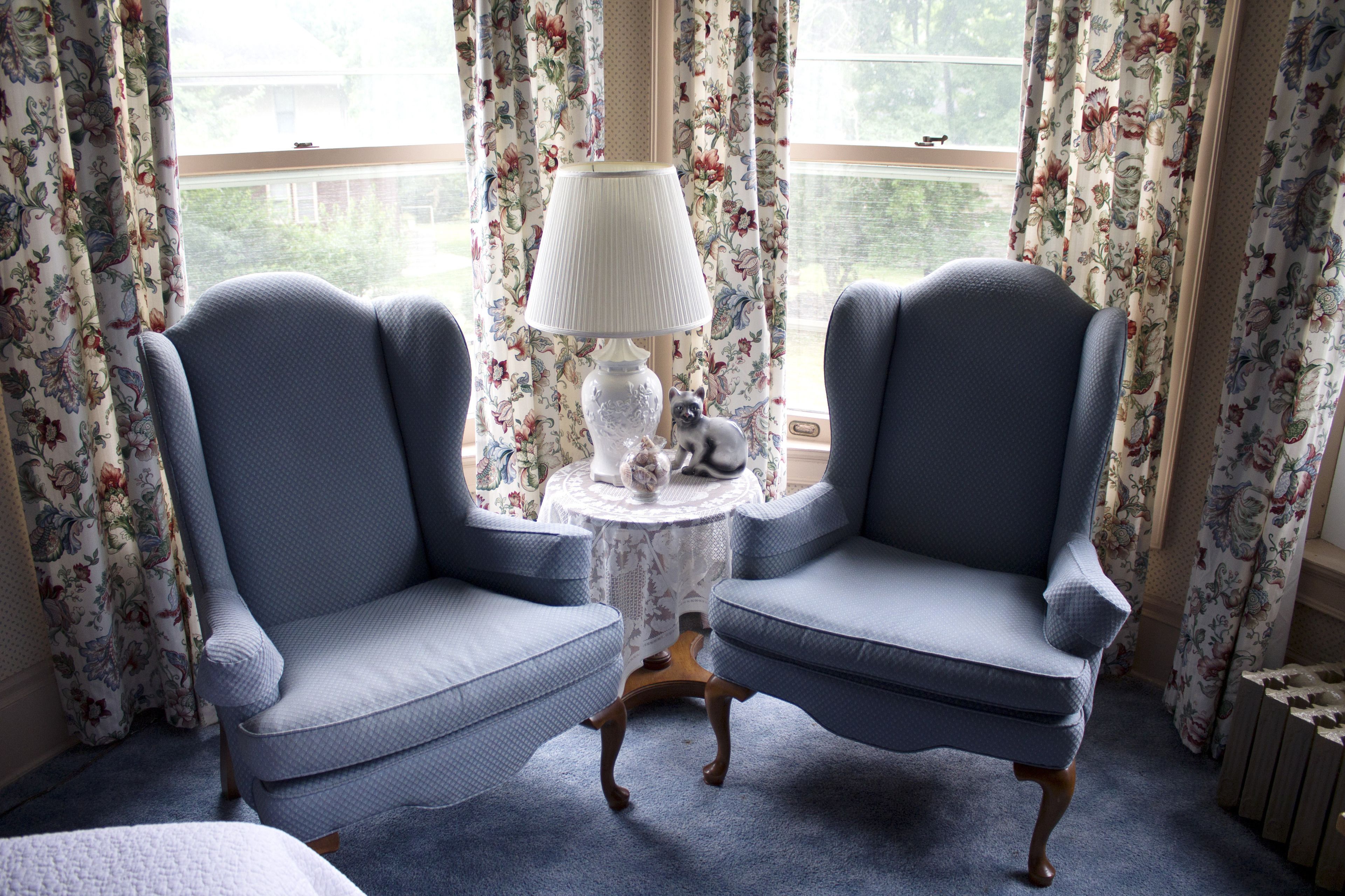 Two blue armchairs facing each other with a small table between them, a lamp, and floral curtains.