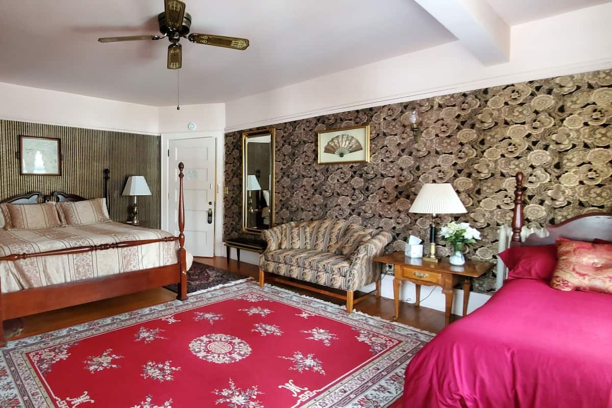 A bedroom with a four-poster bed, a red patterned rug, a wooden dresser, a nightstand with a lamp, and a floral wallpaper design.