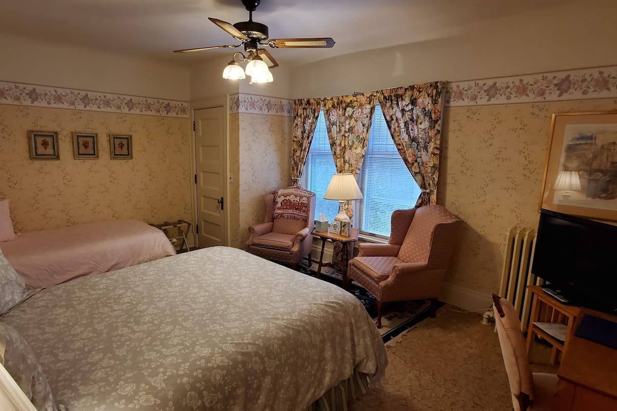A hotel room with two beds, a ceiling fan, a TV, a chair, and a lamp.
