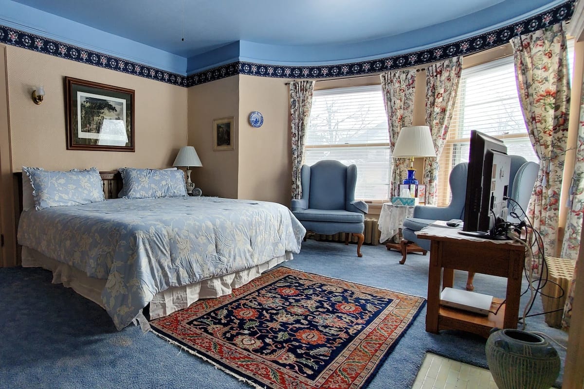 A bedroom with a bed, a chair, a desk, a television, and a window with curtains.
