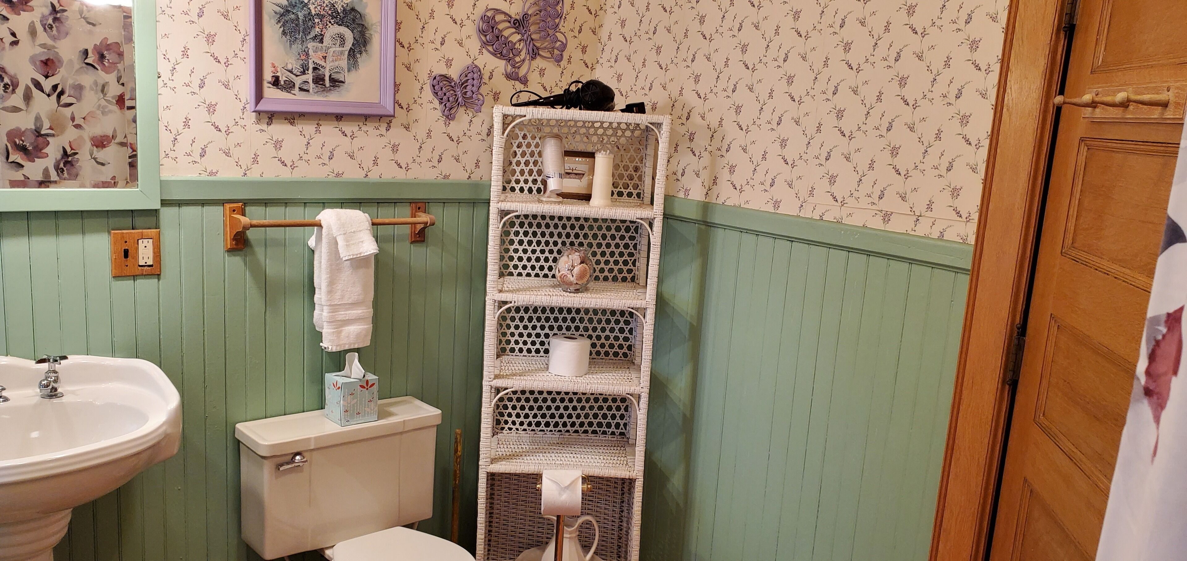 Un baño con lavamanos, inodoro y un estante de mimbre con artículos de aseo.