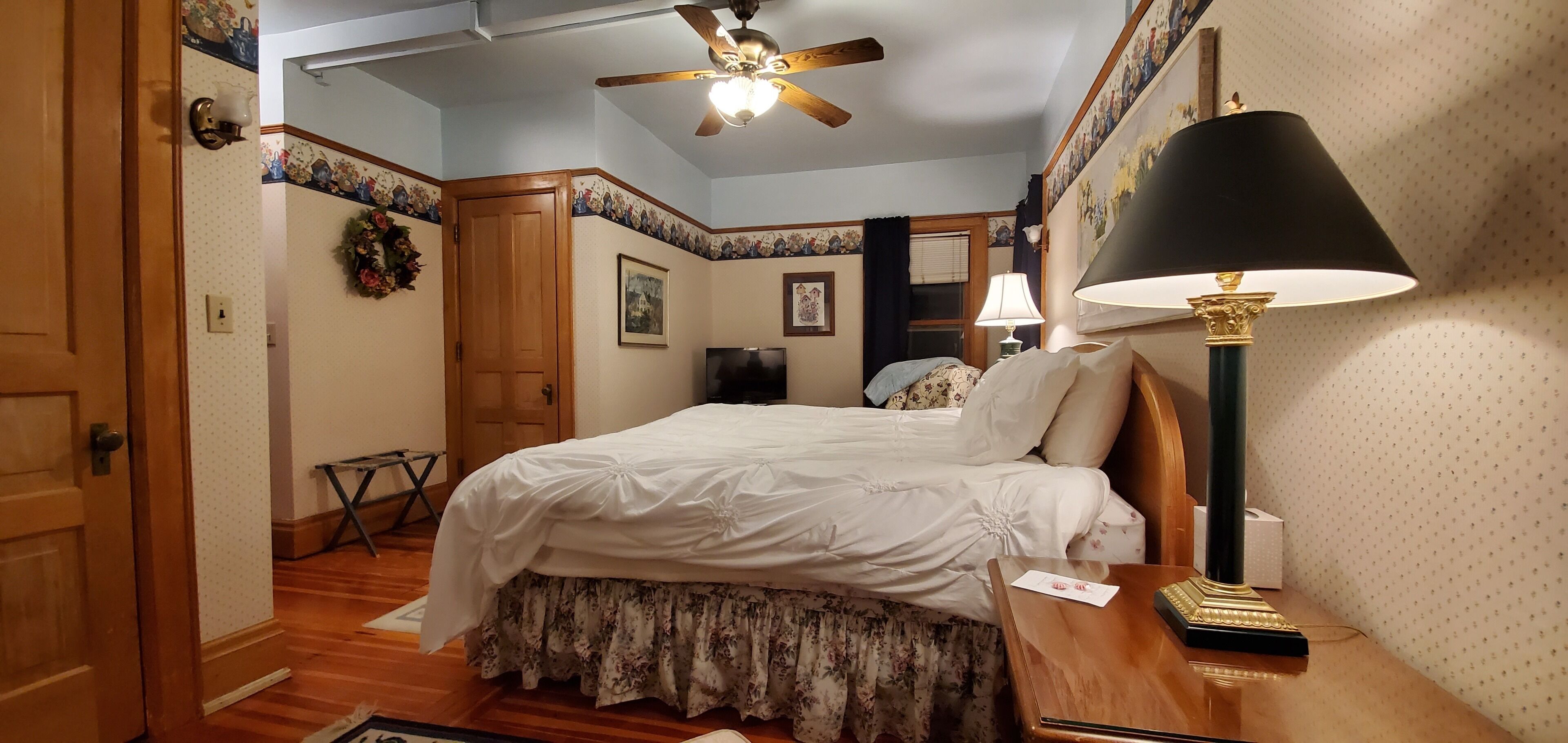 A bedroom with a bed, a ceiling fan, a nightstand with a lamp, and a door.