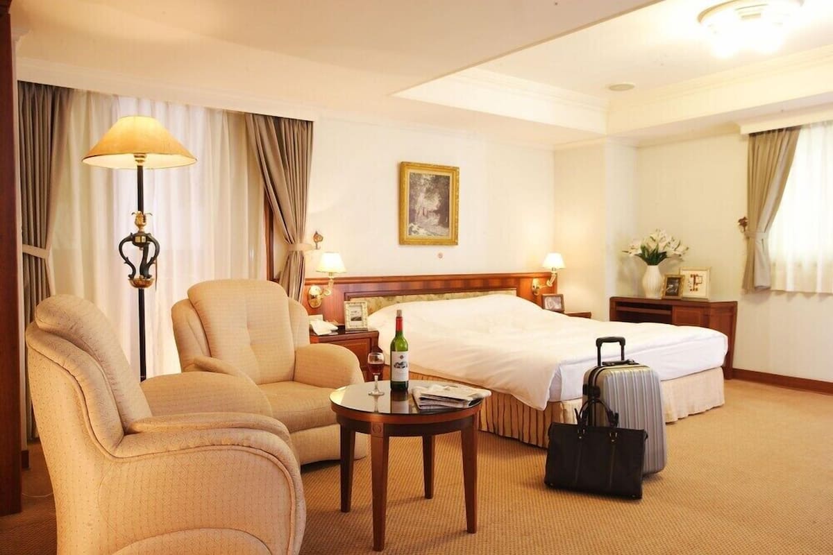 A hotel room with a bed, two armchairs, a small table with a bottle and glass, a suitcase, and a lamp.