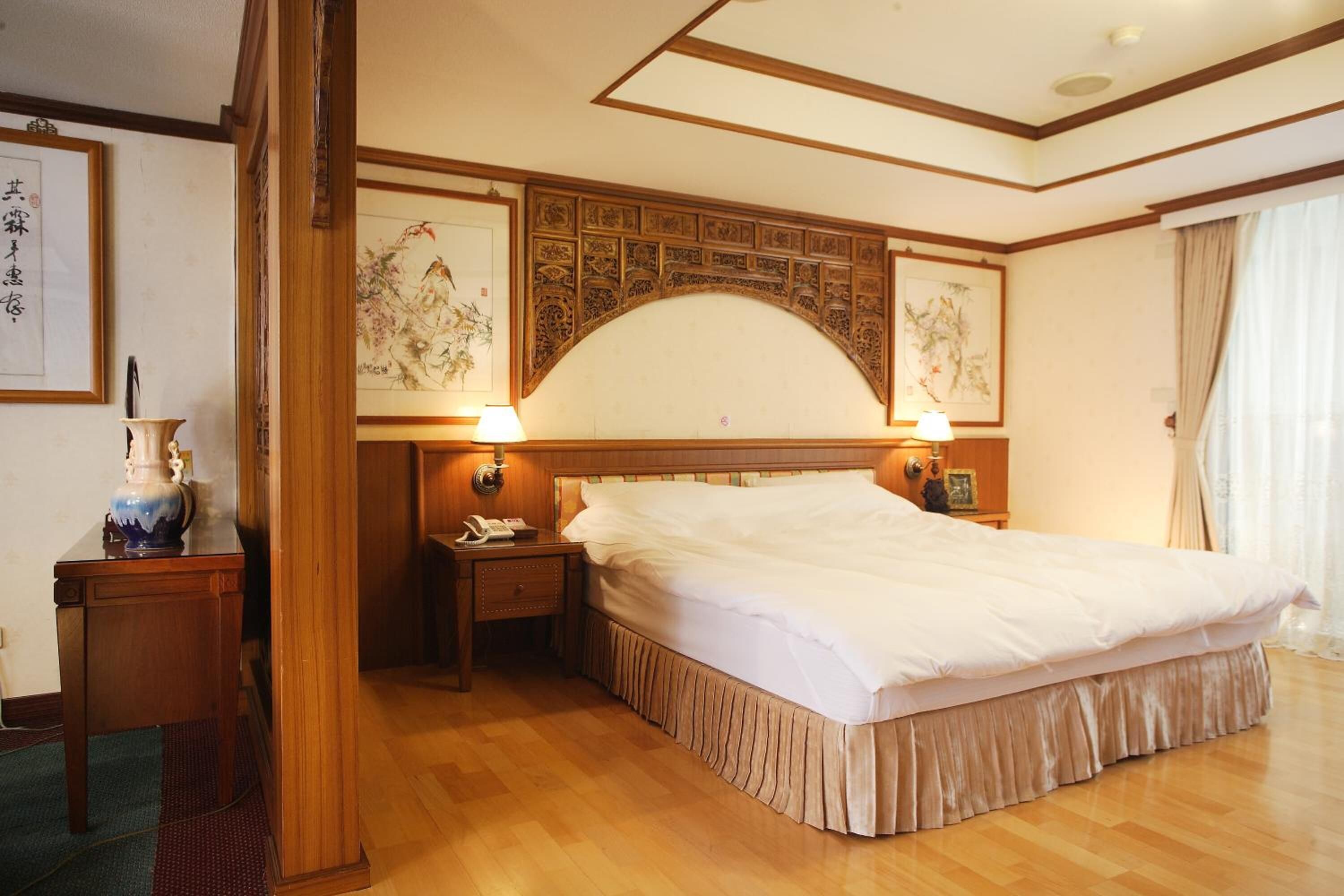 Una habitación de hotel con una cama grande, cabecera de madera, dos mesitas de noche con lámparas, suelo de madera y cuadros en las paredes.