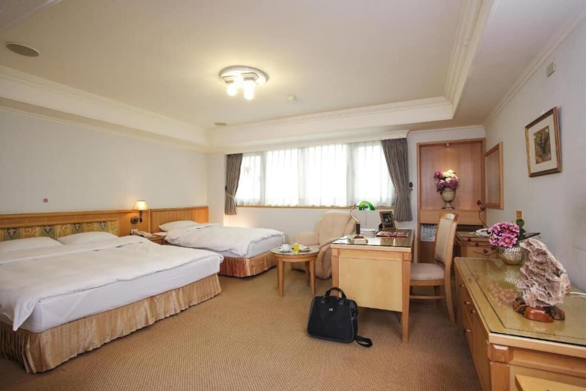 A hotel room with two beds, a desk, a chair, a small table, and a lamp.