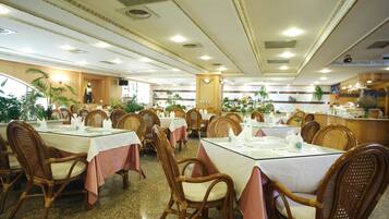 A restaurant with round tables, wicker chairs, and a kitchen in the background.