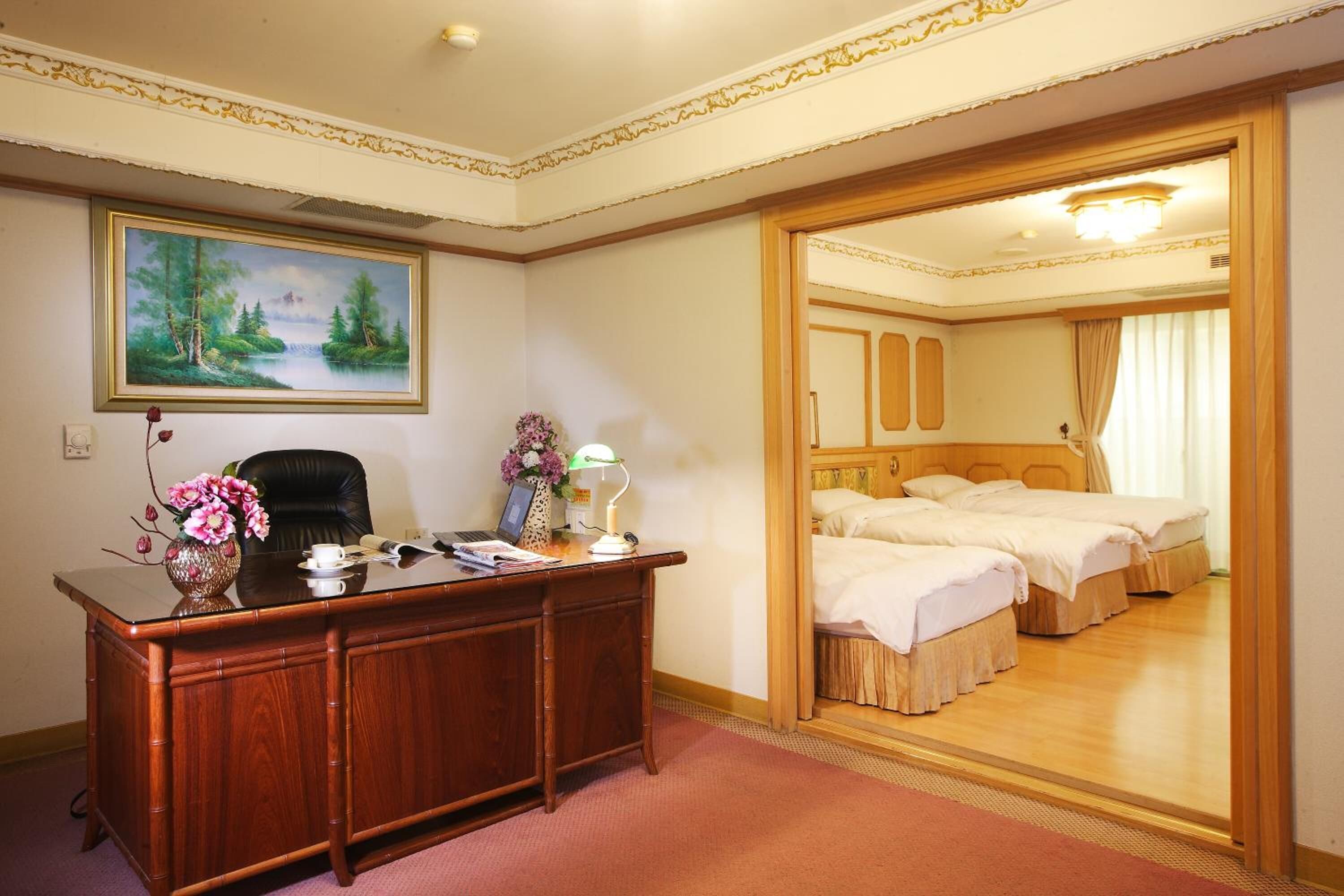 A hotel room with a wooden desk, a painting of a landscape, a bed with two beds, and a mirror.
