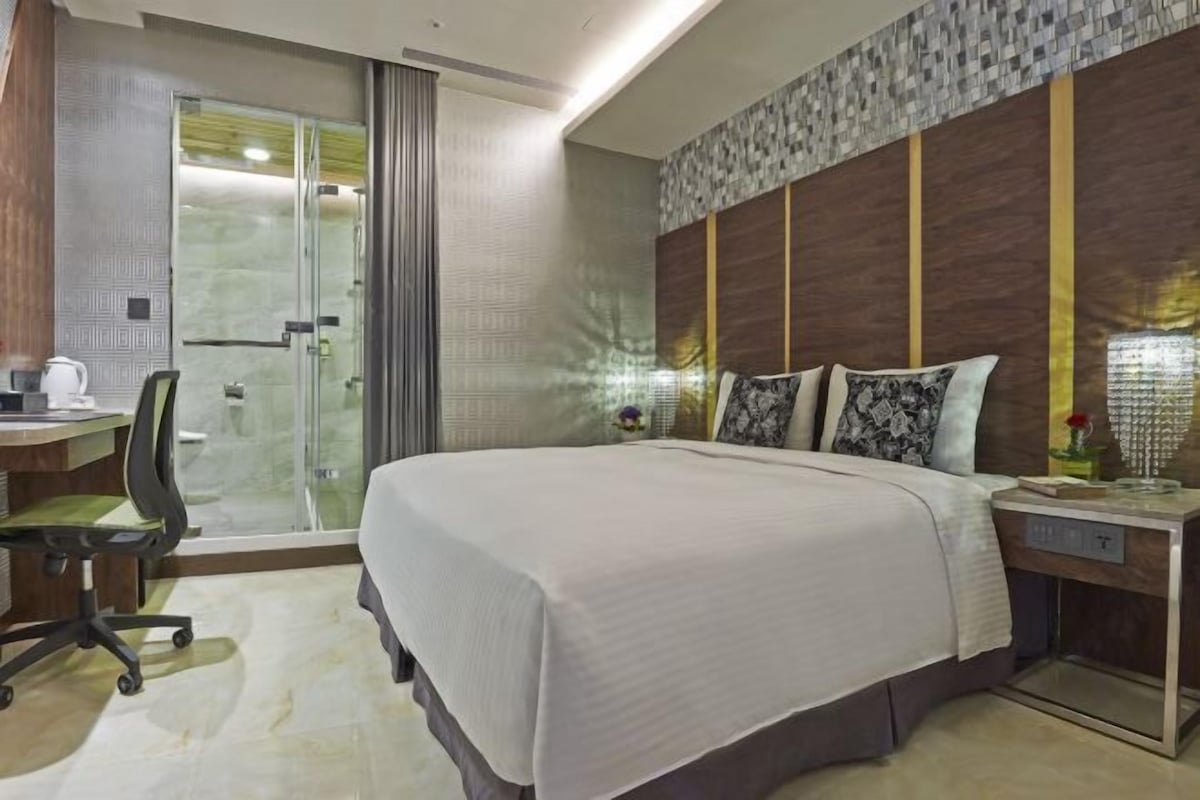 A modern hotel room with a large bed, a desk with a chair, a glass shower enclosure, and a decorative wall panel.