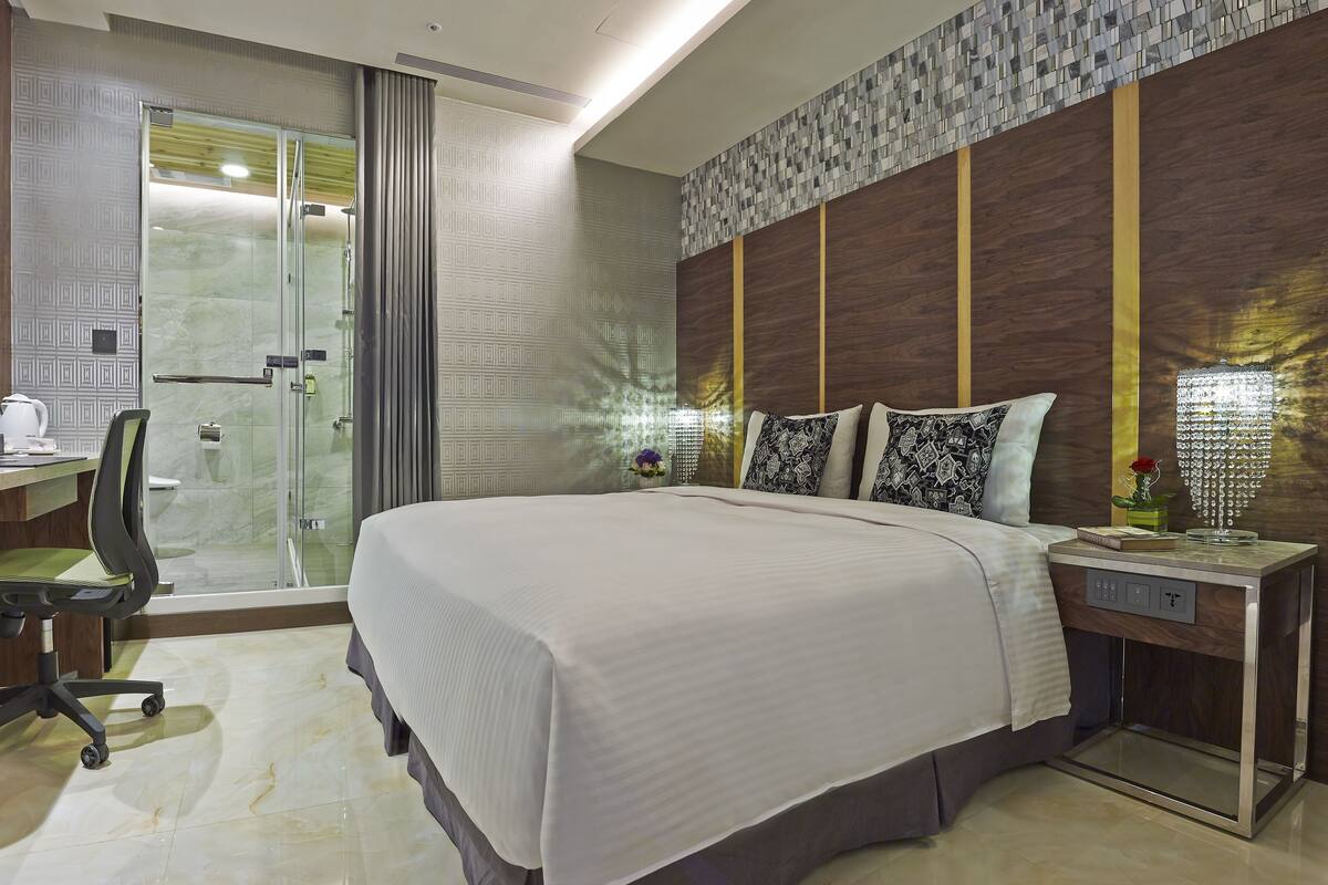 Habitación de hotel con una cama grande, un escritorio con silla, zona de ducha y mesita de noche con lámpara.