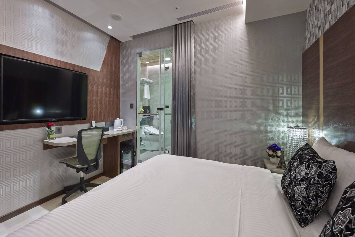 Una habitación de hotel moderna con una cama grande, un escritorio con silla, un televisor de pantalla plana, un baño con ducha de vidrio y un jarrón decorativo con flores.