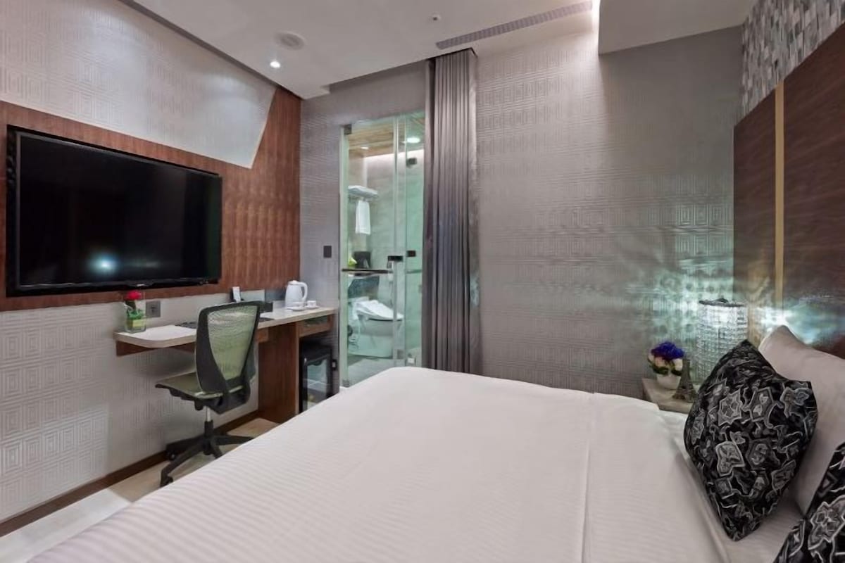 A hotel room with a bed, a TV mounted on the wall, a desk with a chair, and a bathroom visible through a glass door.