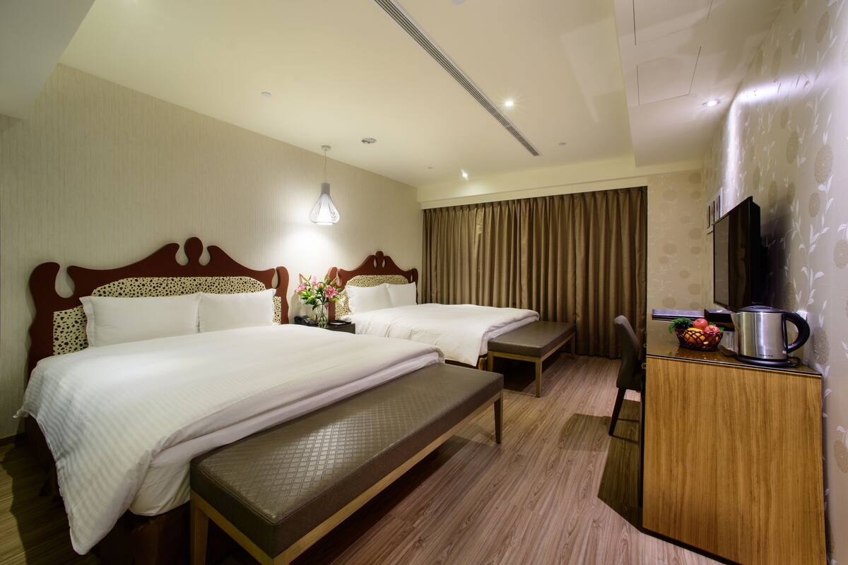 Una habitación de hotel con dos camas, un banco, un escritorio con hervidor y un televisor.